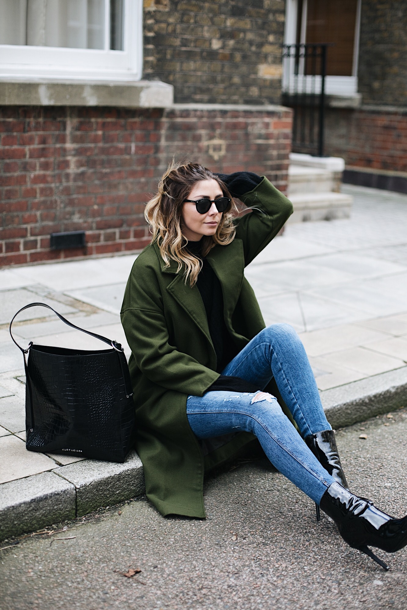 Emma Hill wears khaki coat, black sweater, ripped skinny jeans, large croc bag, black patent vinyl heeled ankle boots, round Rayban sunglasses, chic winter outfit