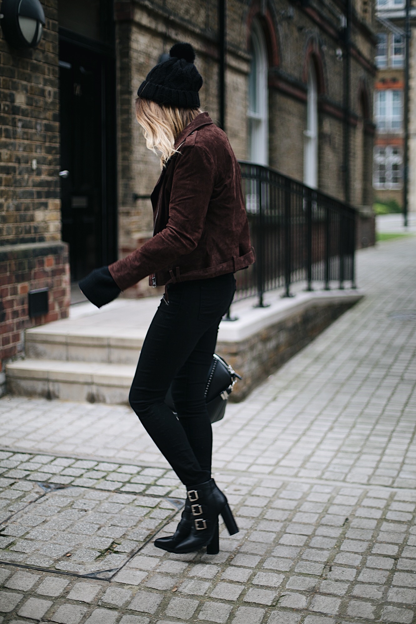 Emma Hill wears brown suede jacket, black skinny jeans, black sweater, knitted bobble hat, buckled heeled ankle boots, studded Givenchy Antigona bag, Round Rayban sunglasses, winter outfit ideas