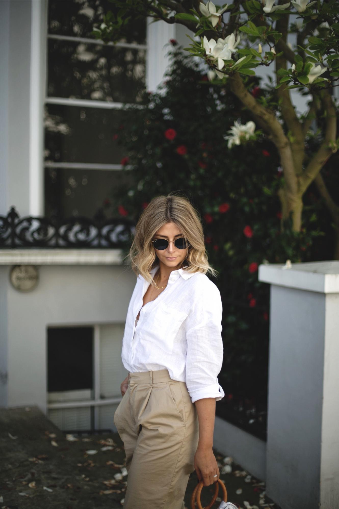 Emma Hill wears white linen shirt, beige high waisted wide leg chino trousers, Simon Miller tan nubuck suede ring handle bonsai bag, scarf tied to bag, chic Spring outfit