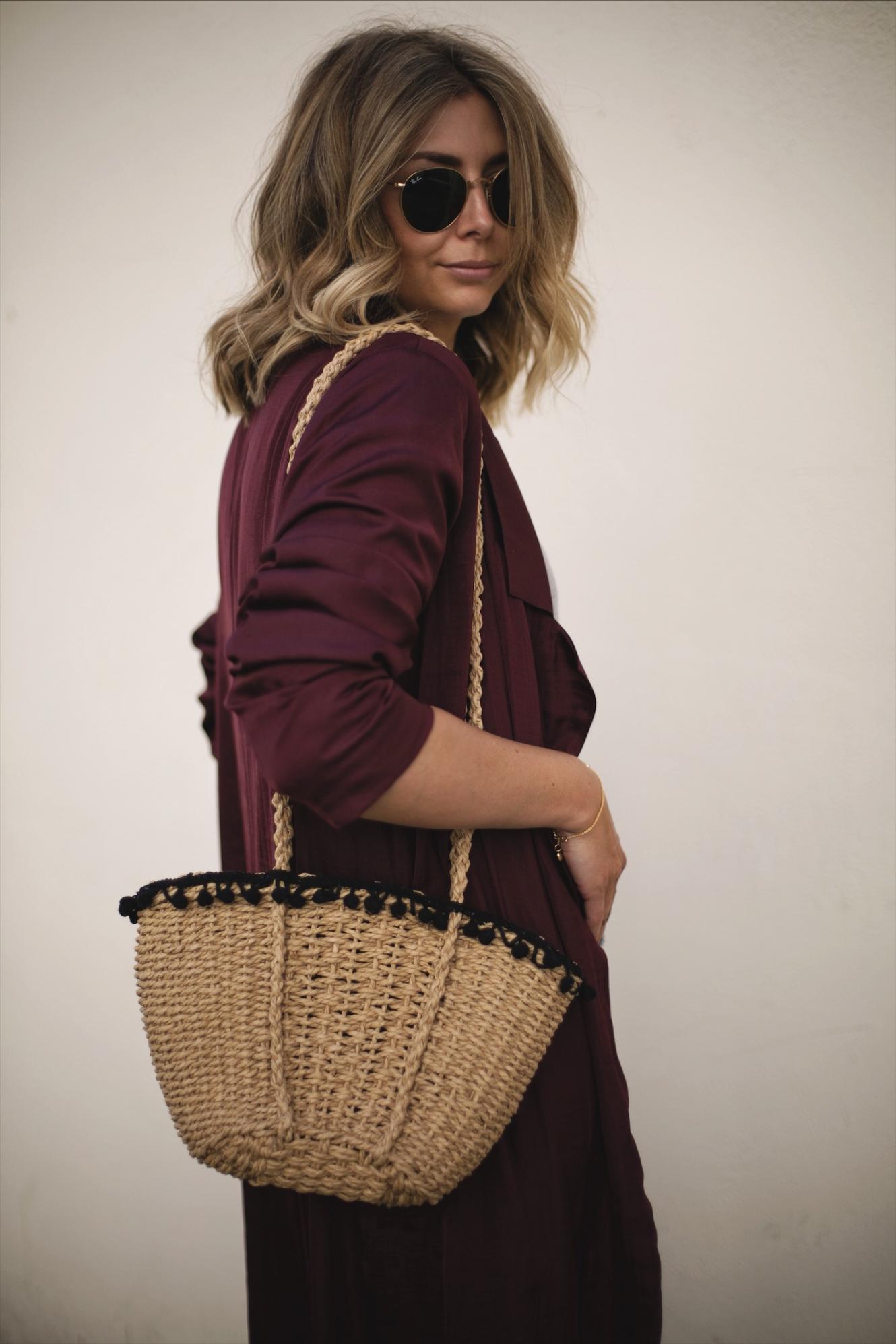 Emma Hill wears wine coloured long silky duster jacket kimono, straw basket bag with pom pom detail, gold Ray-Ban round metal sunglasses, blonde lob hair