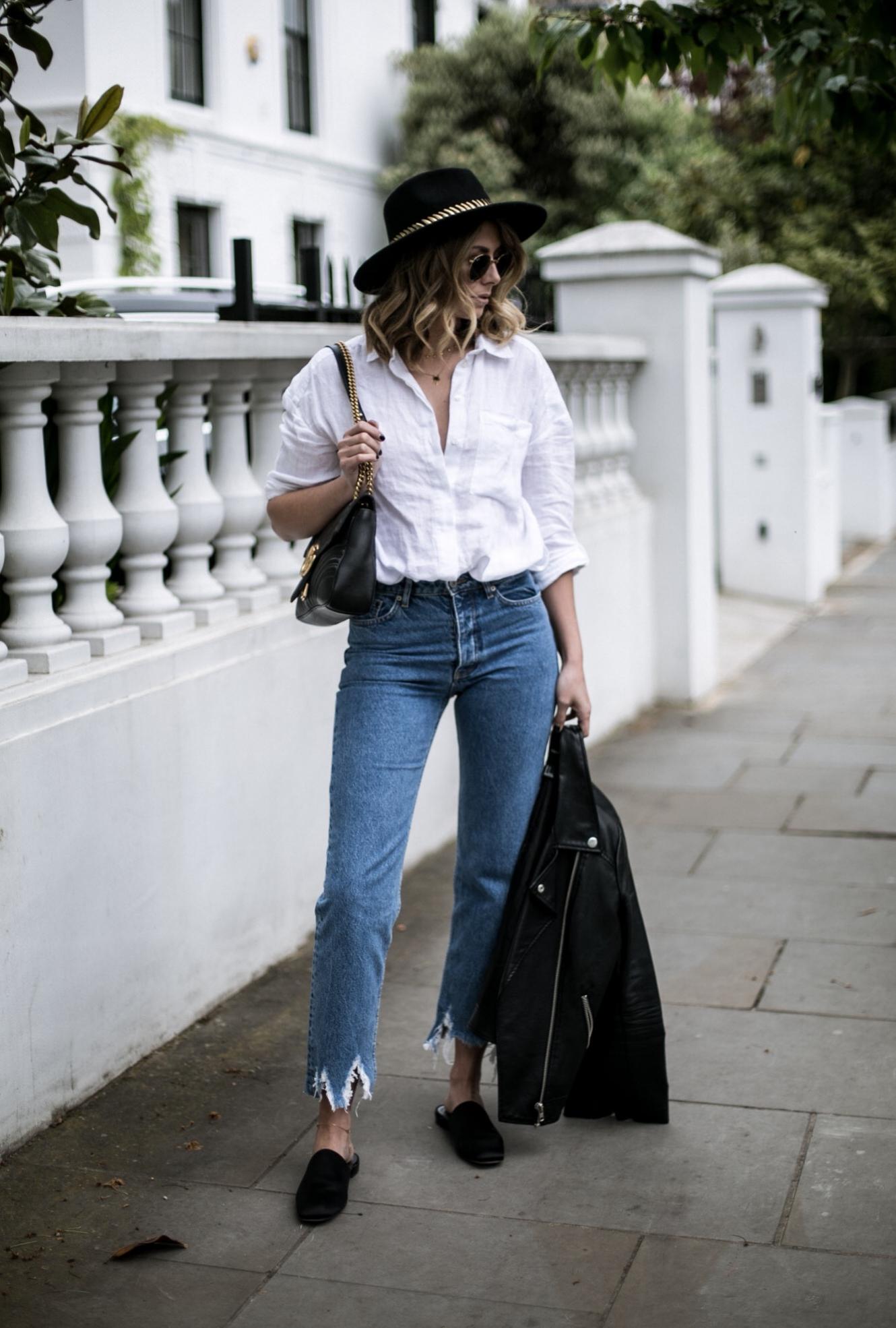 Emma Hill of EJSTYLE wears white linen shirt, distressed hem jeans, black leather biker jacket, fedora with gold band, Gucci Marmont bag, satin mules, casual spring outfit